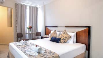 Une chambre d’hôtel avec un grand lit, un bureau avec une chaise, une petite table avec un vase de fleurs et une fenêtre avec des rideaux.