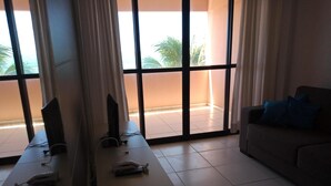 A room with a large window, a sofa with blue cushions, and a table with electronic devices. - Atlântico Hotel (Maceió)