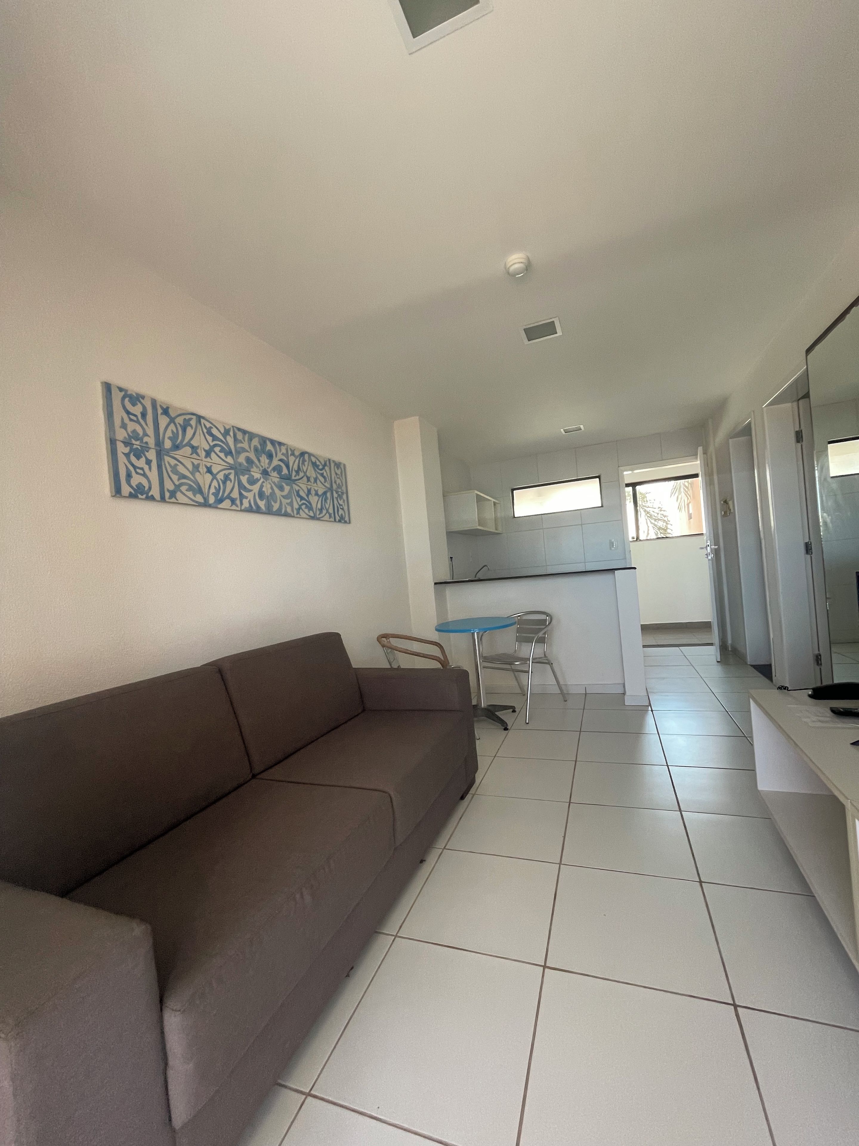 Una sala moderna con un sofá marrón, una mesita redonda y un sillón. Se ve un área de cocina al fondo con armarios y un ventanal.