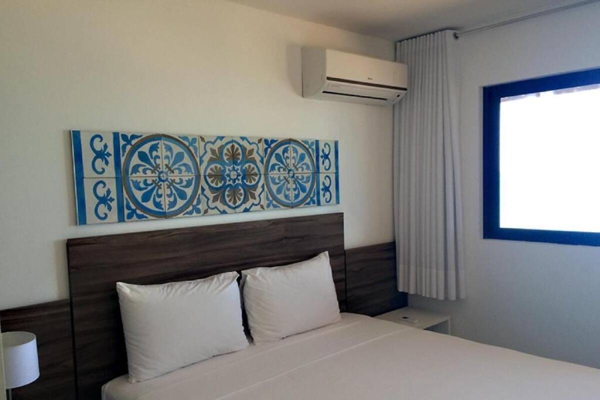 A hotel room with a bed, a nightstand, a wall-mounted air conditioner, and a window with curtains.