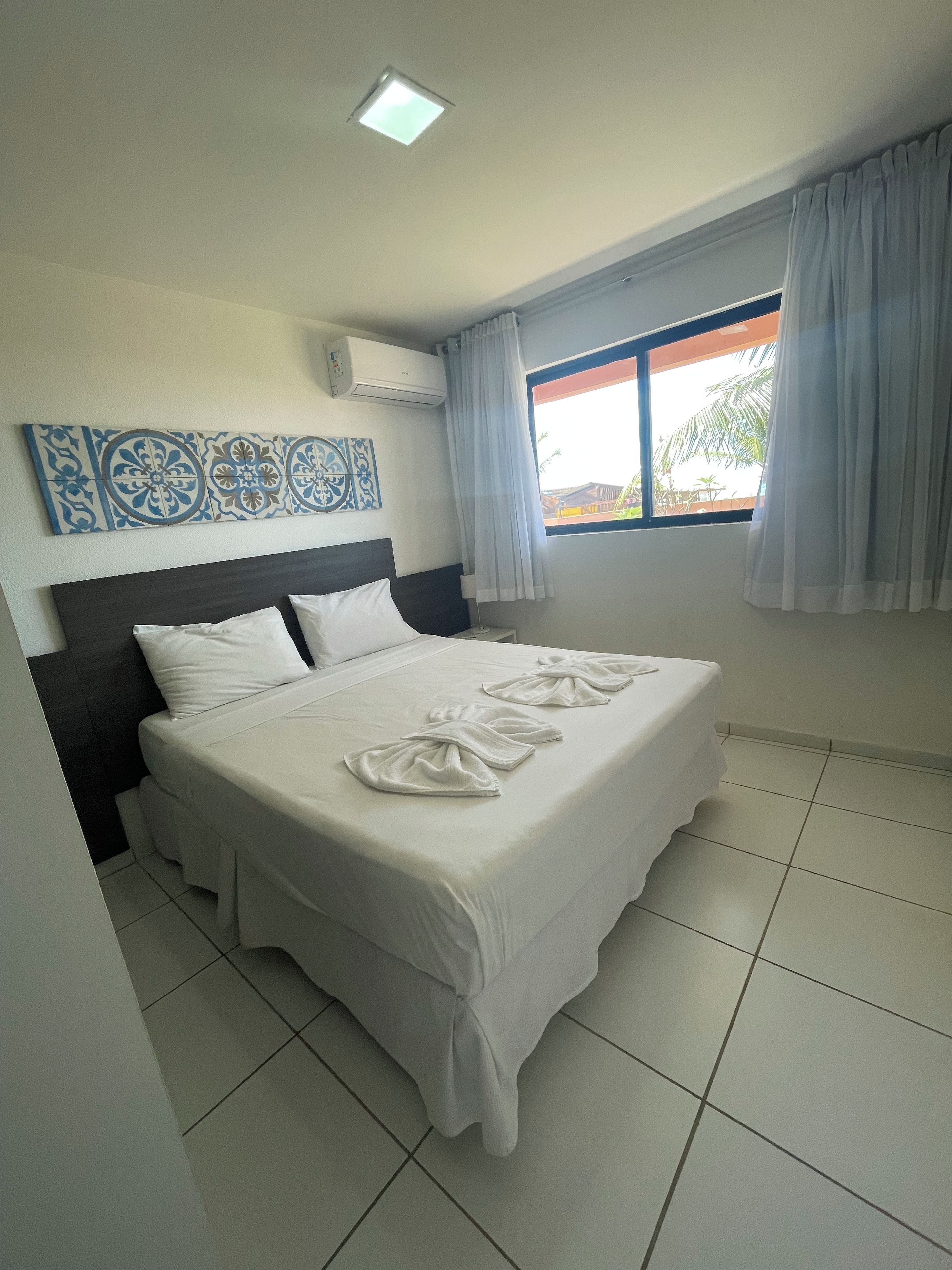 A hotel room with a bed, a window with curtains, a ceiling light, and a wall-mounted air conditioner.