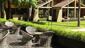 Outdoor wicker furniture arranged on a wooden deck, a wooden building with a sloped roof, and a well-maintained lawn with trimmed bushes. - Grand Admiral Resort & SPA (Irpin)