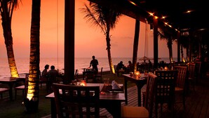 An outdoor dining area with tables and chairs, palm trees, and a view of the ocean at sunset.