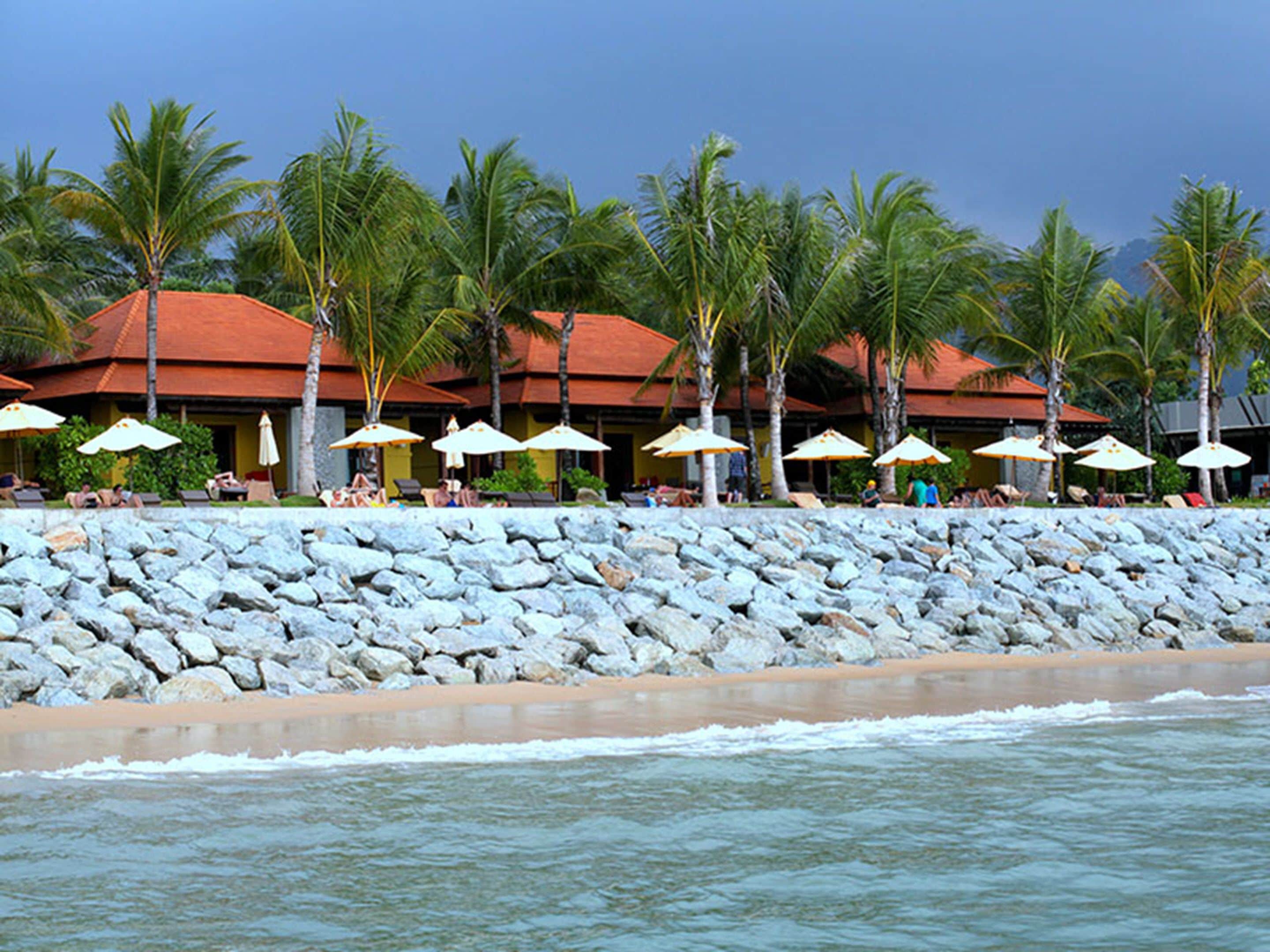 Un resort frente a la playa con muro de piedra, palmeras y sombrillas.