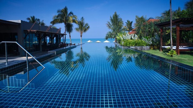 A clear blue infinity pool with a view of the ocean, surrounded by palm trees and a covered seating area.
