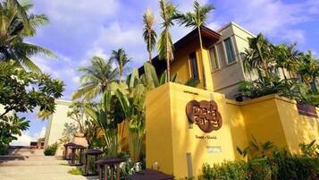 A hotel entrance with a yellow facade, palm trees, and outdoor seating.