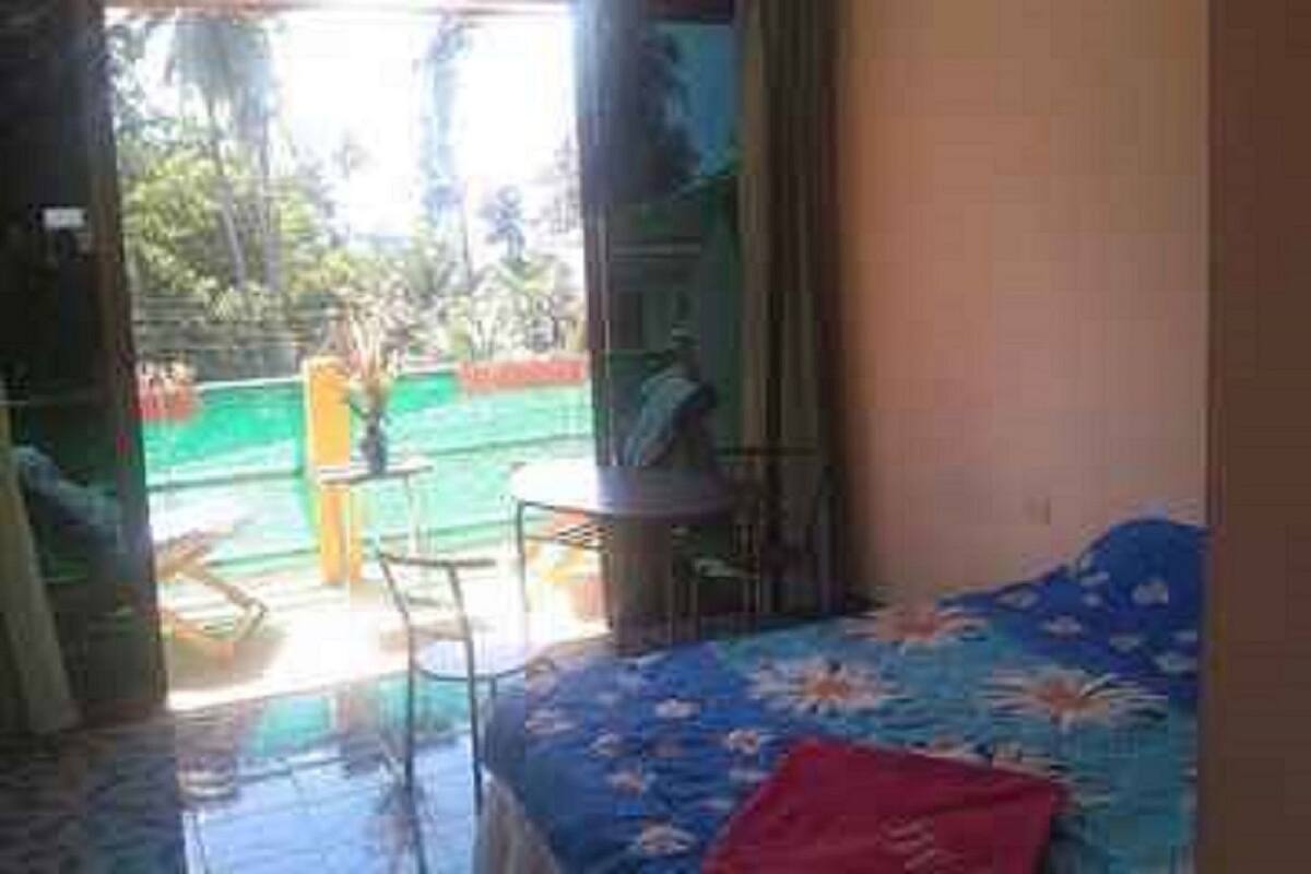 A room with a bed, a small table, and a chair. There is a view of a green outdoor area with a yellow chair.