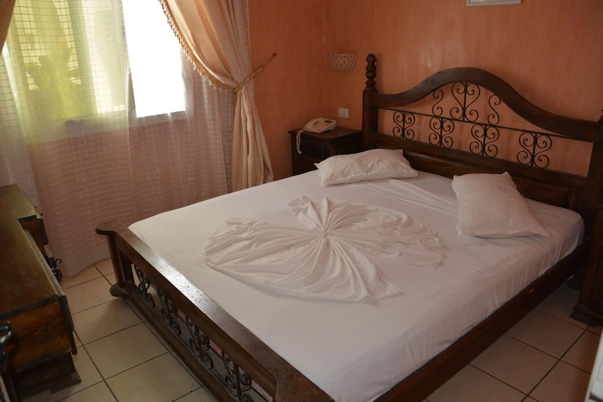 A neatly made bed with a decorative white cloth, a wooden headboard with intricate designs, a bedside table with a telephone, and a window with curtains.
