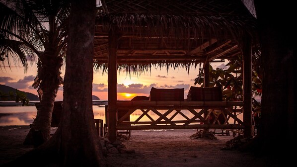 A beach hut with a thatched roof, wooden benches, and a view of the sunset over the water.