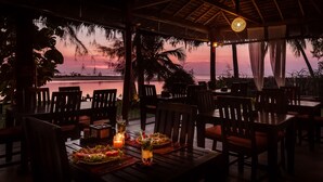 Un área de comedor al aire libre con mesas y sillas de madera, vista al atardecer y una masa de agua tranquila de fondo.