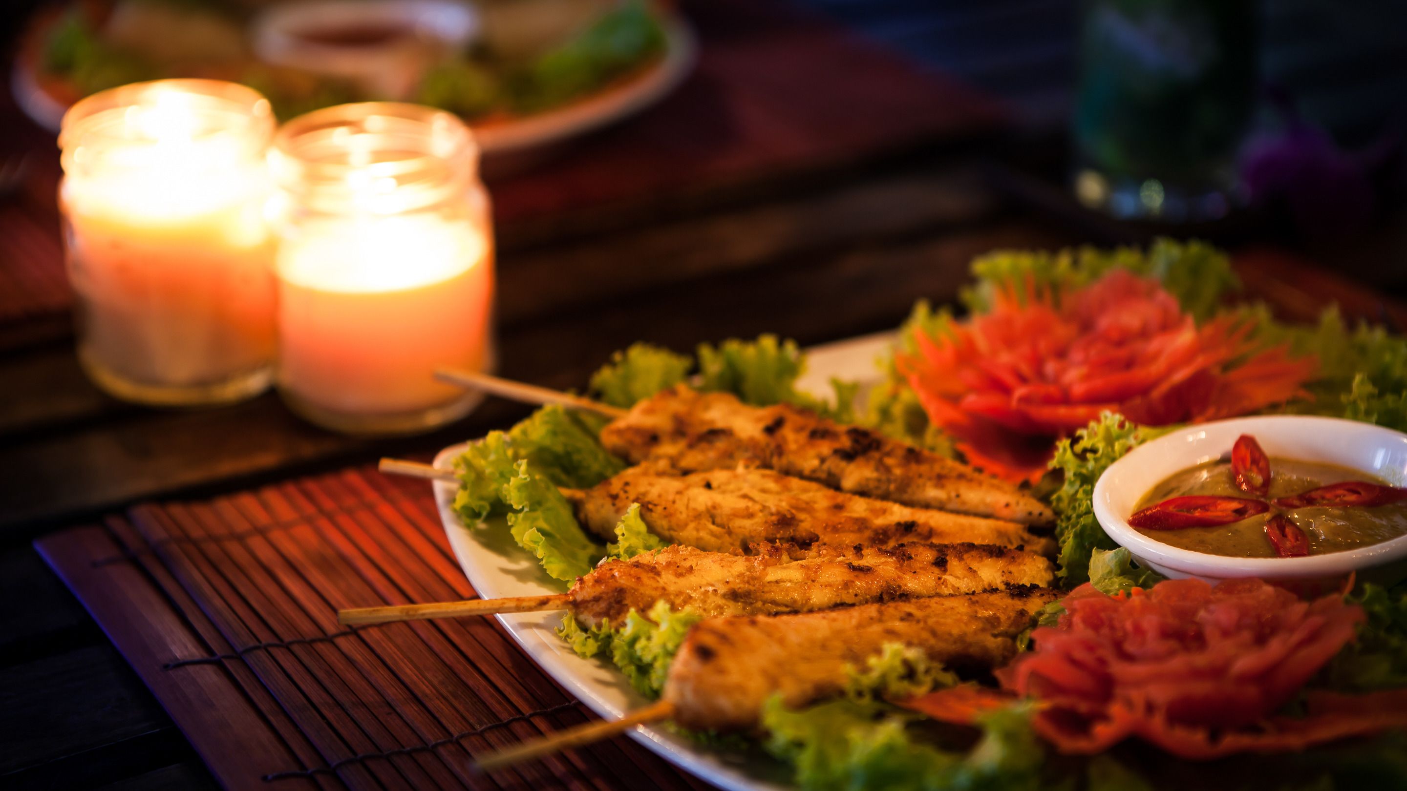 A plate of grilled chicken skewers with lettuce and a small bowl of sauce, set on a table with candles.