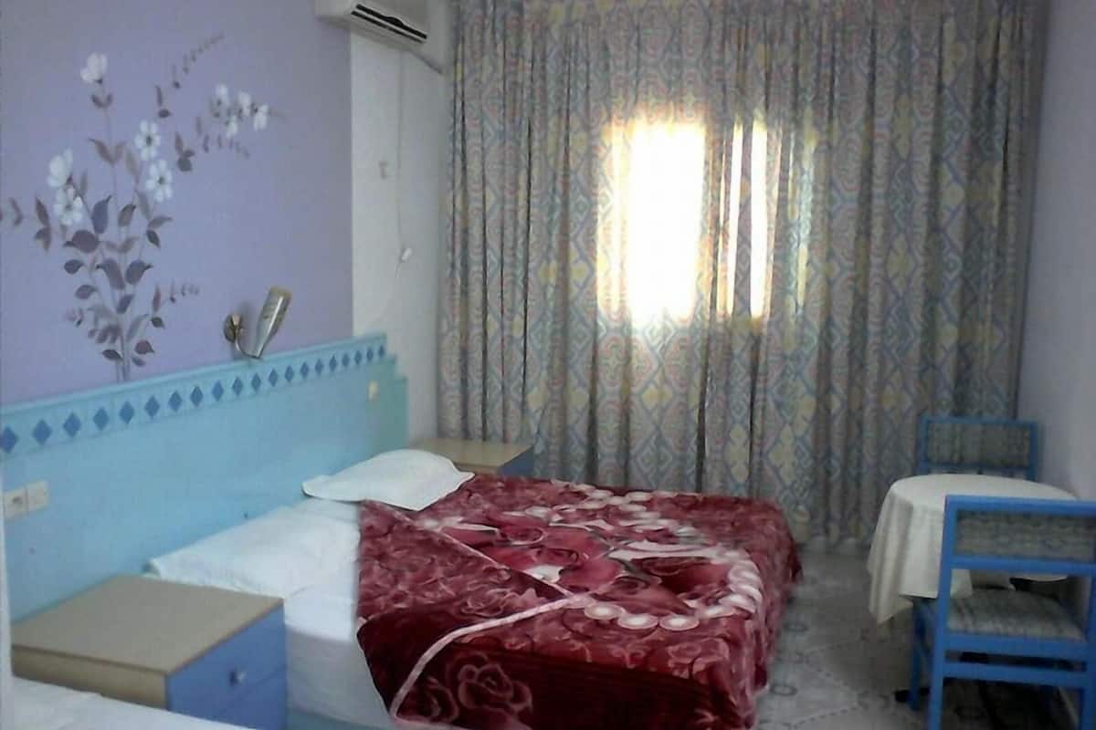 Una habitación con una cama, una mesita pequeña y una silla. Hay una ventana con cortinas estampadas.
