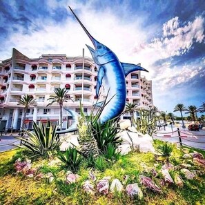 A large blue fish sculpture in front of a multi-story hotel with balconies and palm trees. - Houria House Sable d'Or (Mahdia)
