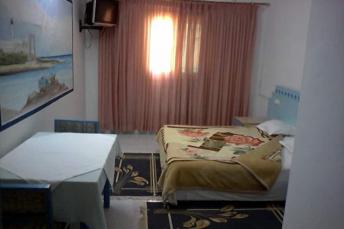 Un dormitorio con cama, escritorio y silla. Hay un cuadro en la pared, una ventana con cortinas y un televisor montado.