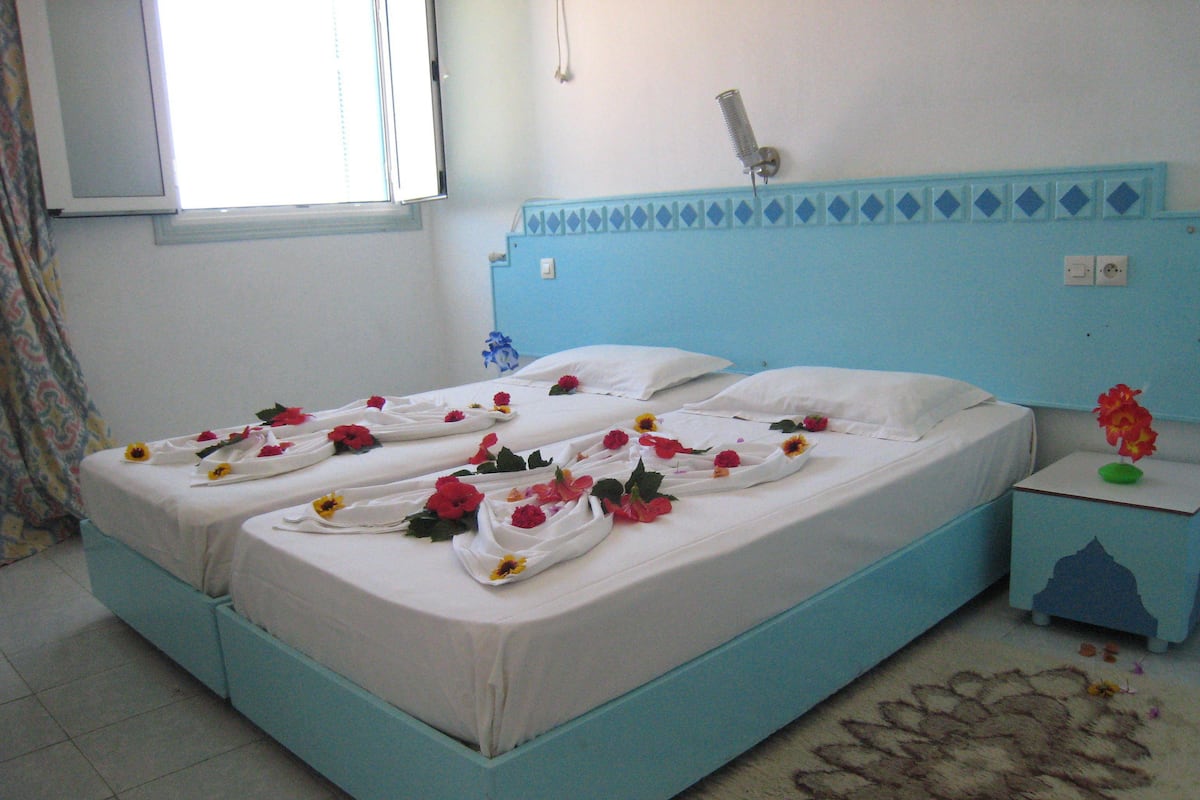 Una cama con sábanas blancas y flores rojas, un cabecero azul, una mesita de noche con un jarrón y una alfombra estampada.
