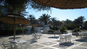 An outdoor patio with white plastic tables and chairs, surrounded by palm trees and thatched umbrellas. - Houria House Sable d'Or (Mahdia)