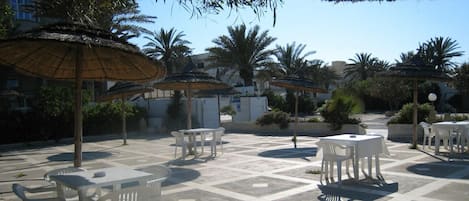 Un patio al aire libre con mesas y sillas de plástico blanco, rodeado de palmeras y sombrillas de paja.