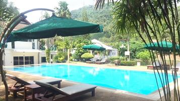 A swimming pool with lounge chairs and umbrellas, surrounded by greenery and a building in the background.