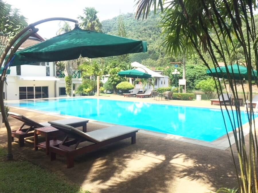 A swimming pool with lounge chairs and umbrellas, surrounded by greenery and a building in the background.