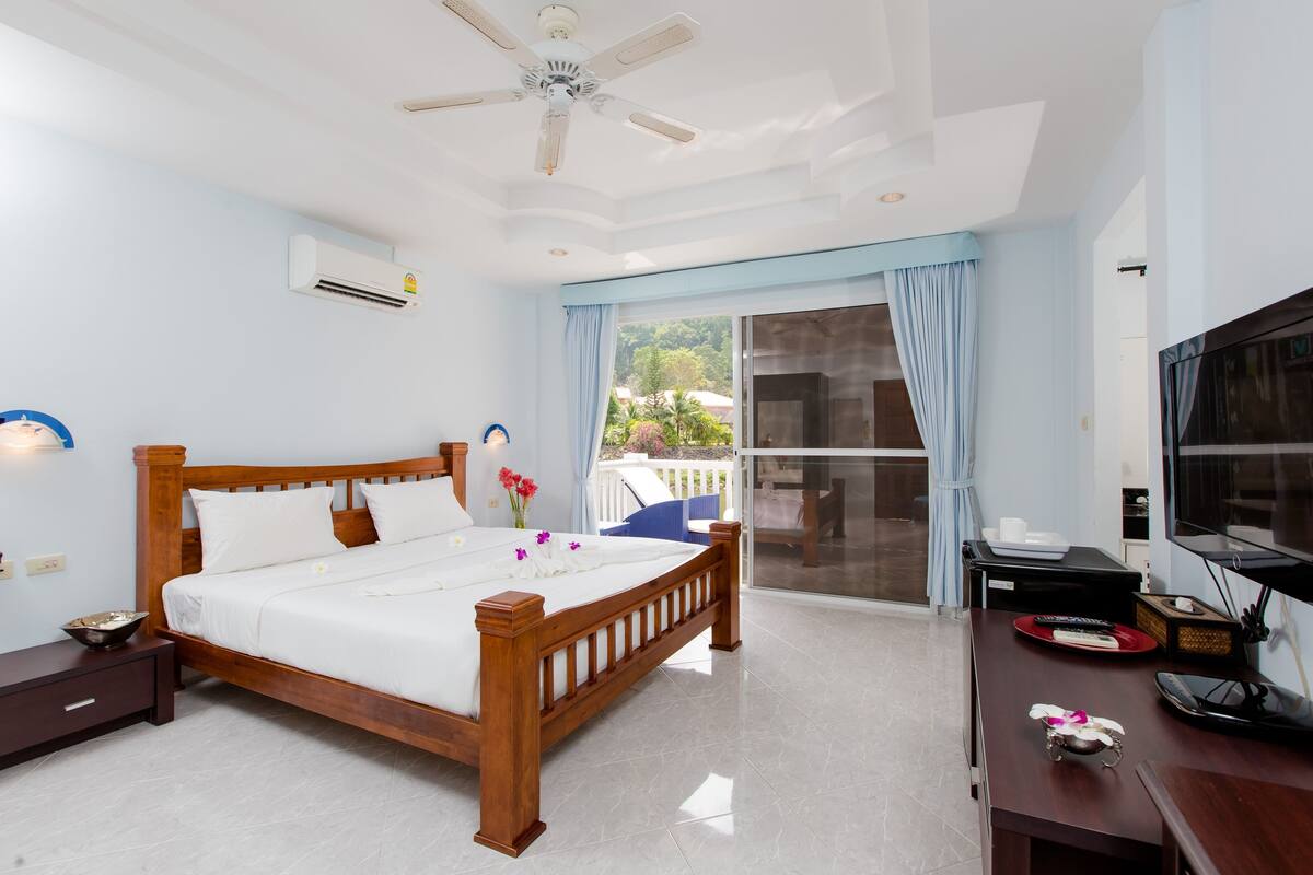 A hotel room with a wooden bed, a ceiling fan, an air conditioning unit, a TV, and a balcony with a view of the outdoors.