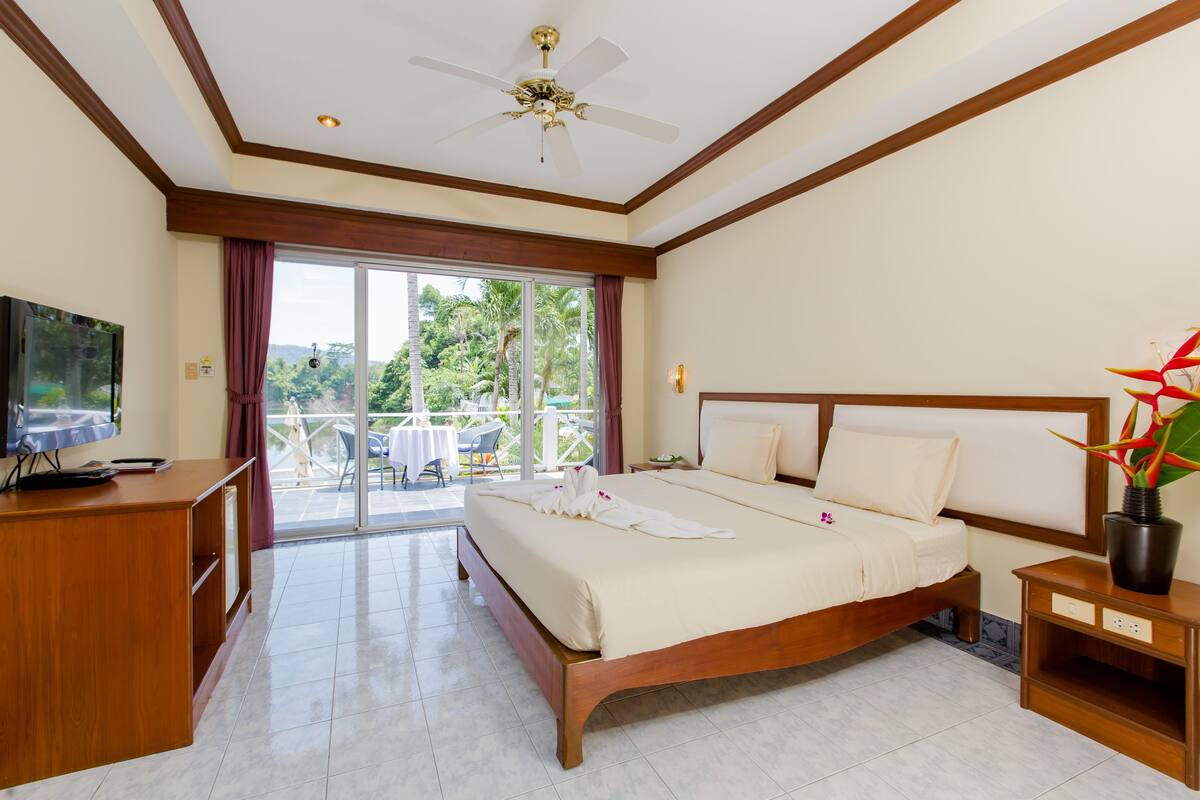 A hotel room with a large bed, a desk, a TV, and a balcony with a view of greenery.