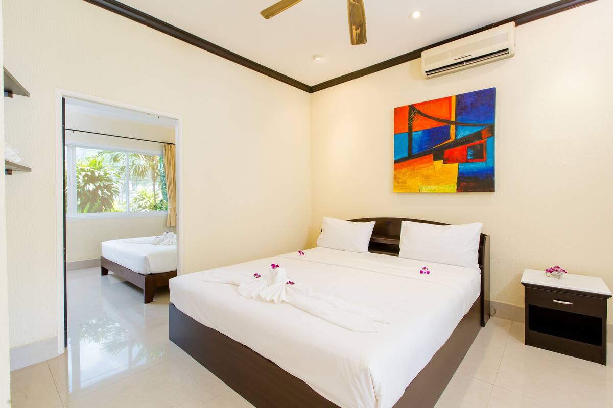 A hotel room with a large bed, a bedside table, an air conditioning unit, and a painting on the wall.