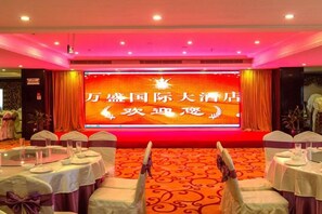 Un salón de banquetes con mesas dispuestas para un evento formal, un escenario con una gran pantalla y un suelo alfombrado.