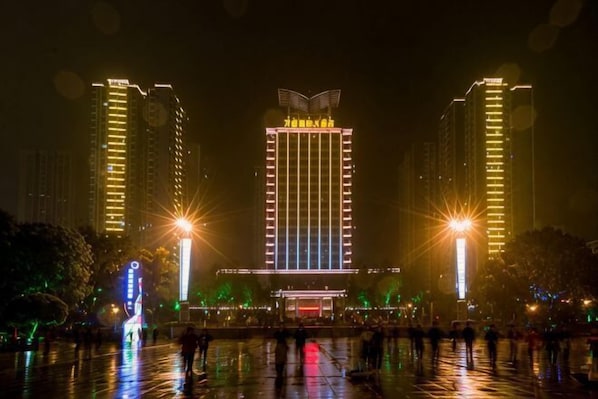 Un edificio de varios pisos con una entrada central, rodeado por una plaza húmeda que refleja luces.