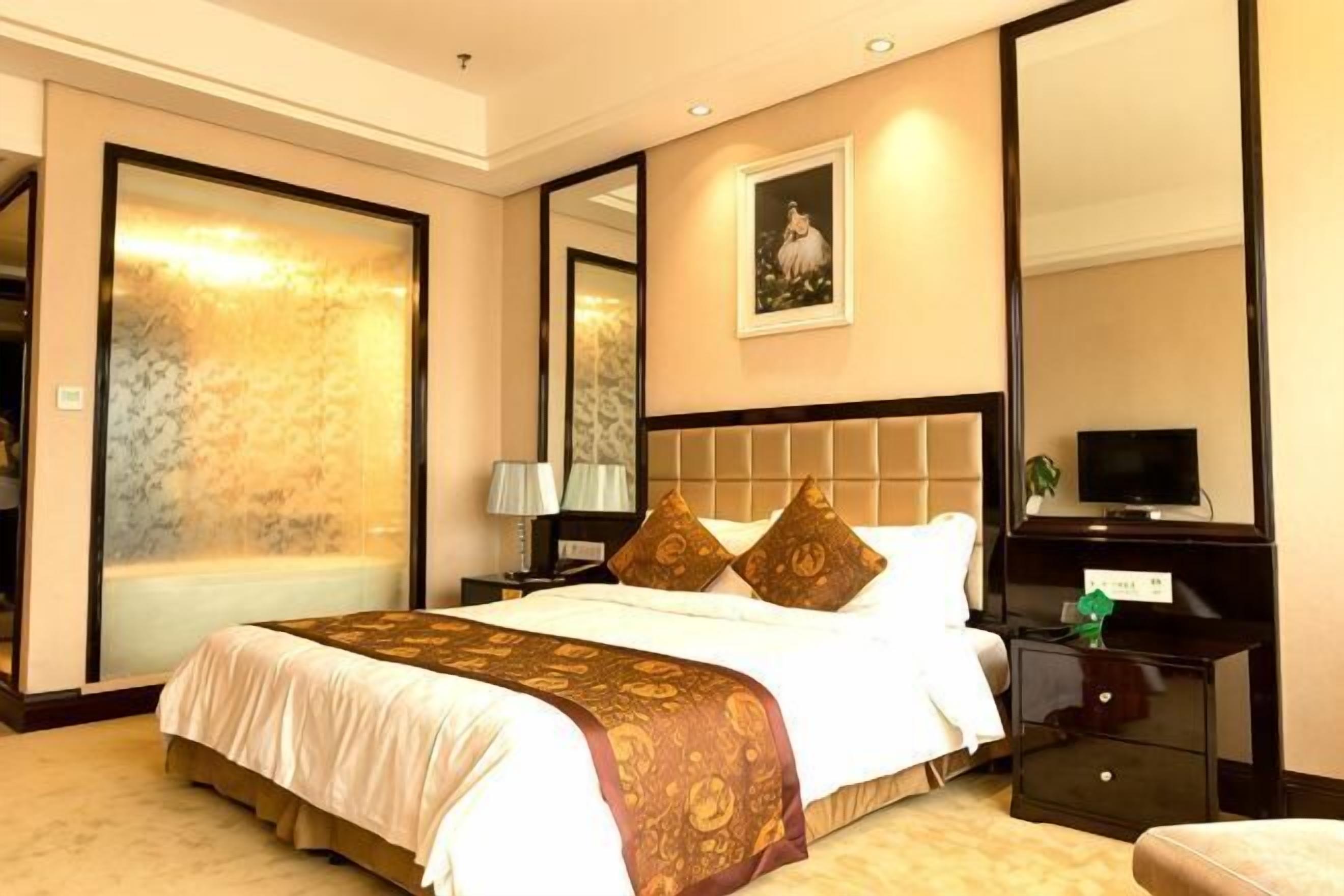 A hotel room with a large bed, a nightstand with a lamp, a television, and a mirror.