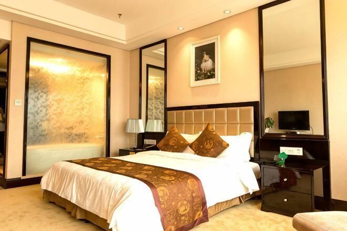 Quarto de hotel com uma cama grande, mesa de cabeceira com abajur, televisão e espelho.