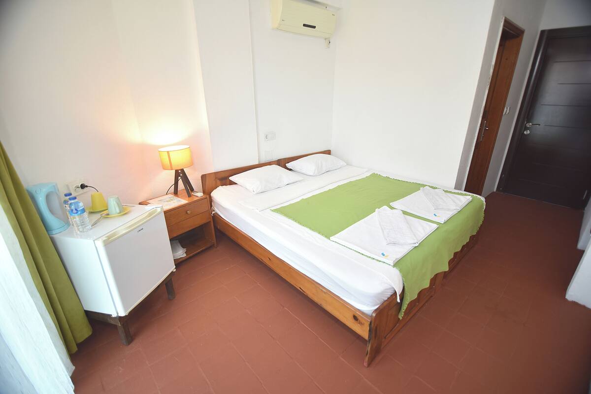 Una habitación pequeña y limpia con una cama individual, una mesita de noche con lámpara, un escritorio pequeño y una puerta.