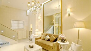 Una sala de estar moderna con un sofá color beige, una mesa de centro de vidrio y una lámpara de araña. Hay un espejo grande en la pared y una escalera que conduce a otro nivel.