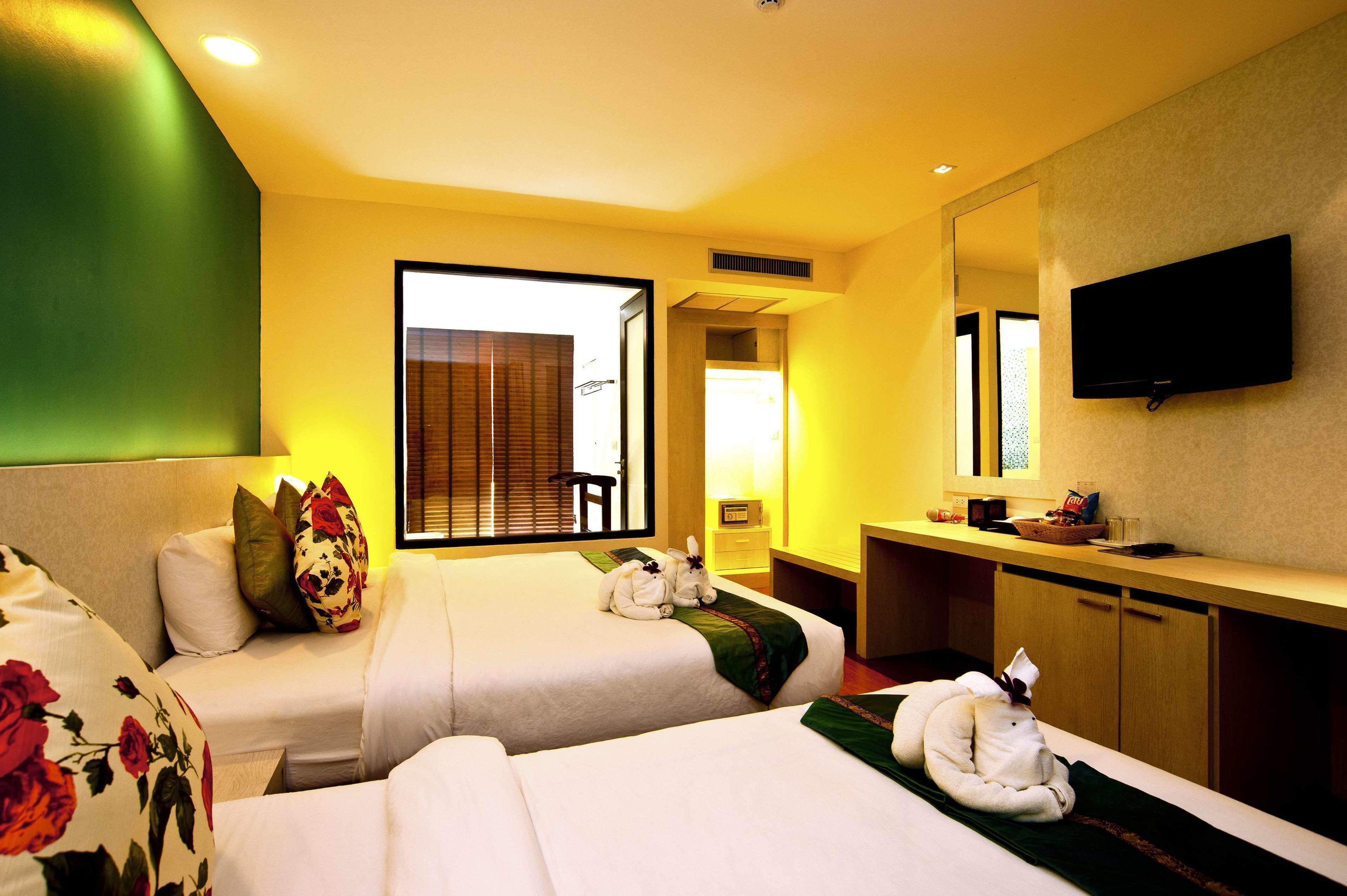 Une chambre d’hôtel avec deux lits, un coin salon avec un fauteuil, un téléviseur fixé au mur et une fenêtre avec des stores.