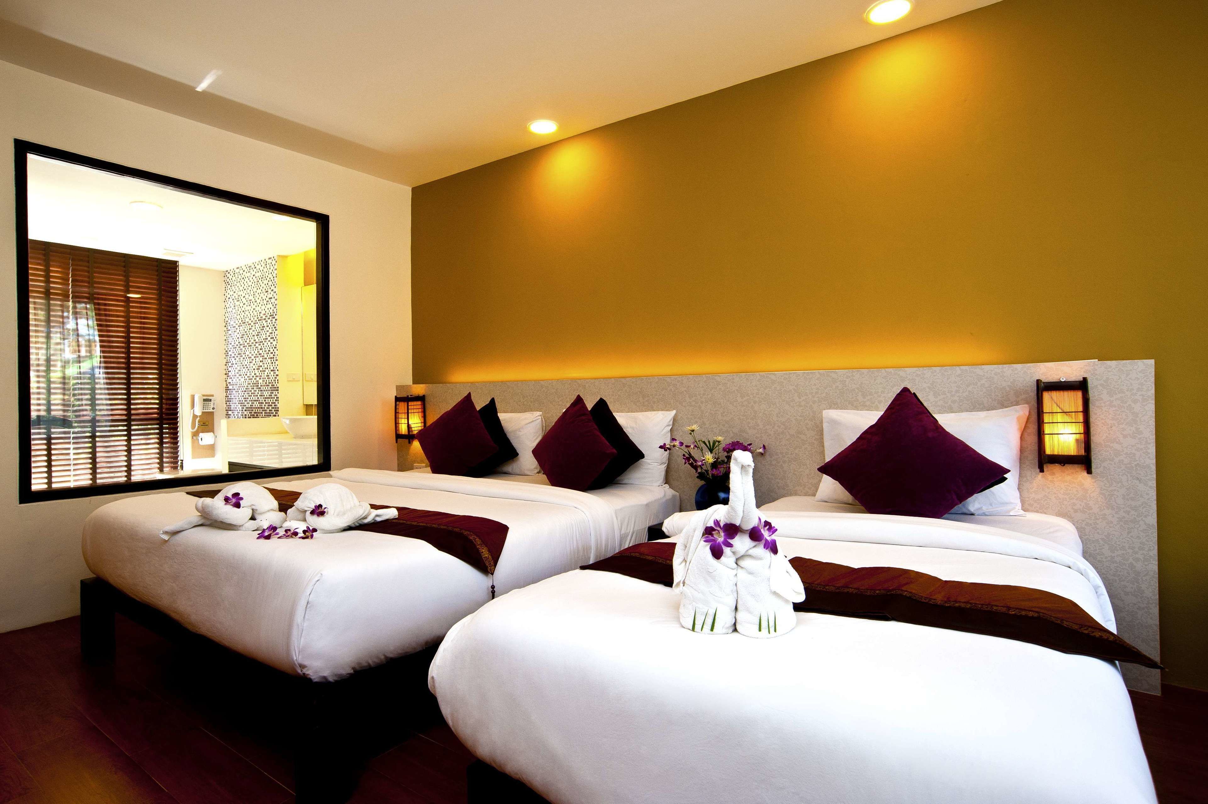Quarto de hotel com duas camas, cada uma com lençóis brancos e almofadas roxas. Há toalhas decorativas nas camas e um vaso com flores.