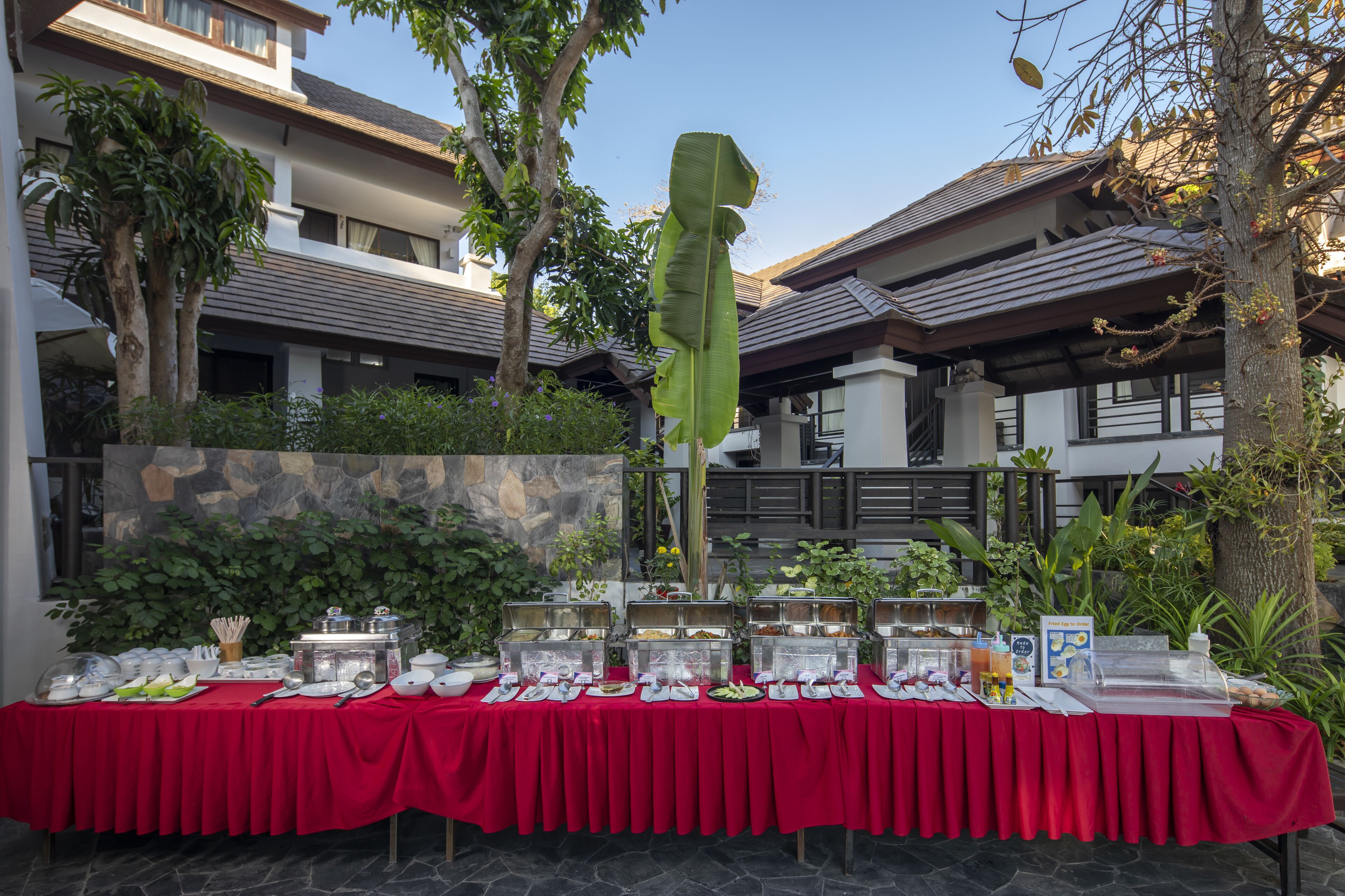 Una mesa de buffet con diversos alimentos y utensilios, montada al aire libre con mantel rojo.