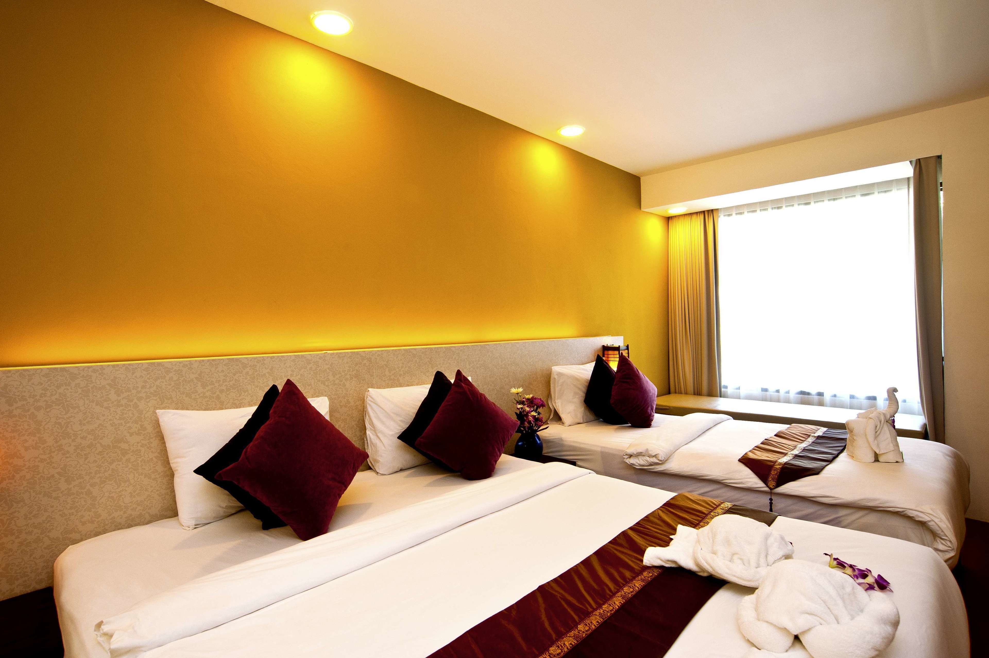 A hotel room with two beds, each with white linens and dark red pillows. There is a small table with a vase of flowers and a towel on one bed. The room has a beige headboard and a large window with curtains.