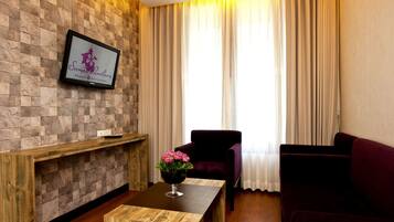 A room with a stone wall, a flat-screen TV, a wooden table with a vase of flowers, a purple armchair, and a sofa.