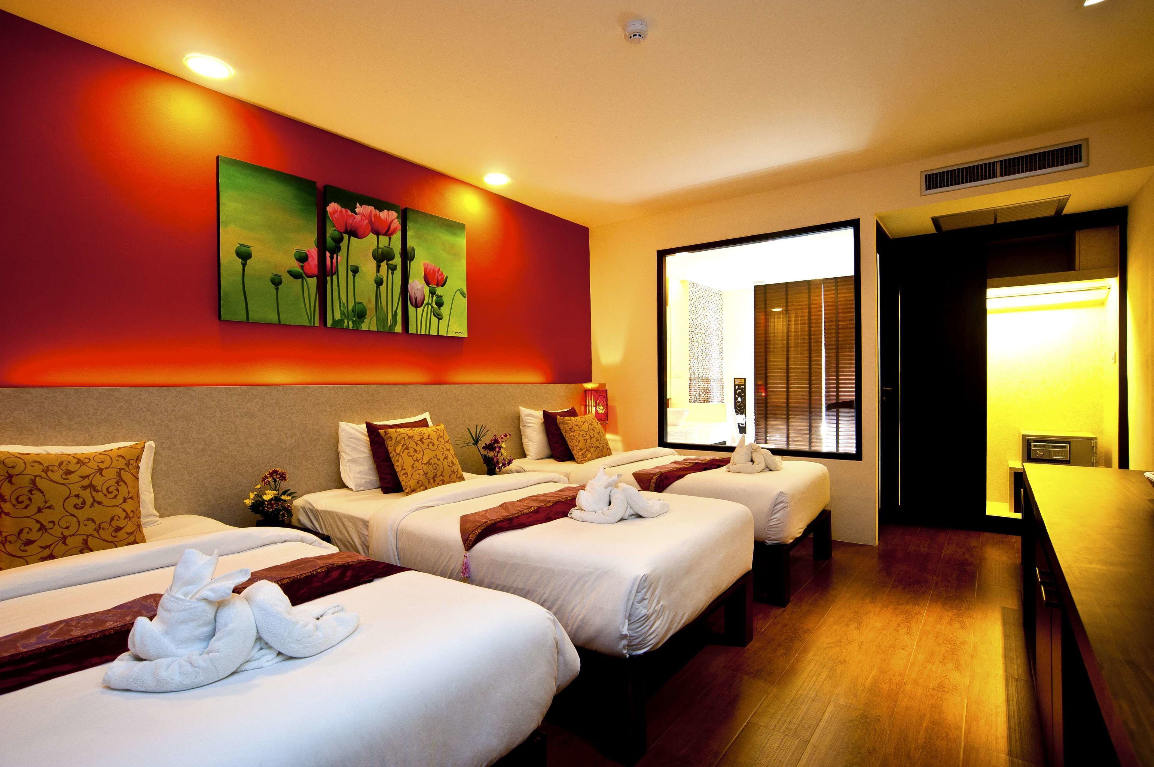 A hotel room with three beds, each with white linens and decorative pillows. There is a large painting of flowers on the wall, and a window providing natural light.