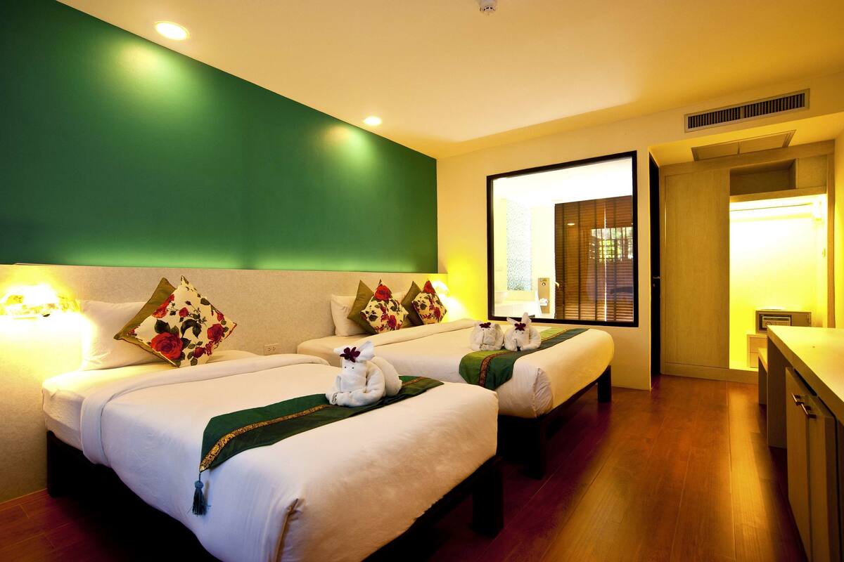A hotel room with two beds, each with a green throw blanket and decorative pillows.