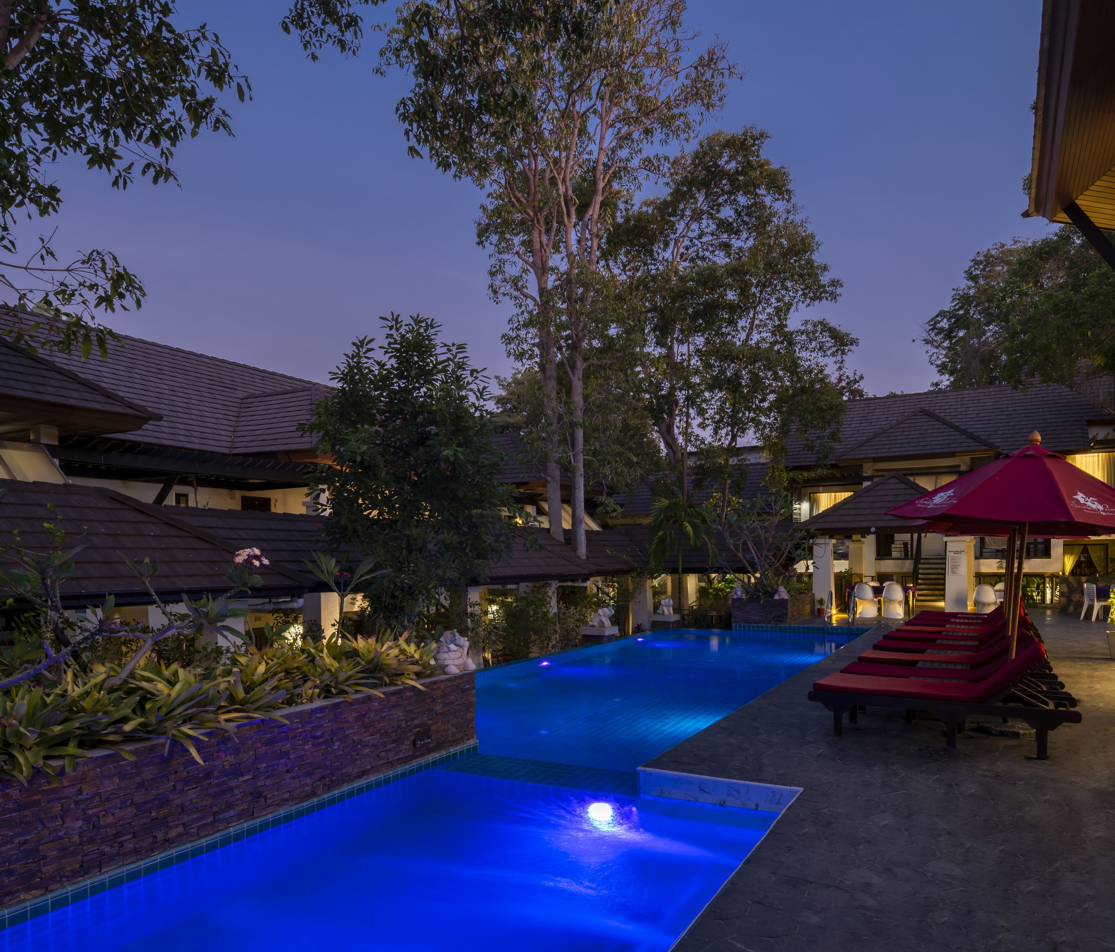 A swimming pool area with lounge chairs and umbrellas, surrounded by trees and buildings with tiled roofs.