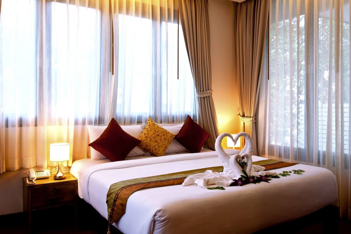 A hotel room with a large bed, two pillows, a bedside table with a lamp, and a heart-shaped decoration on the bed.