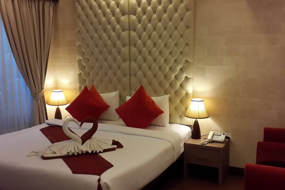 A hotel room with a tufted headboard, a bed with white bedding and red pillows, a heart-shaped decoration on the bed, two bedside lamps, a nightstand with a phone, and a red armchair.