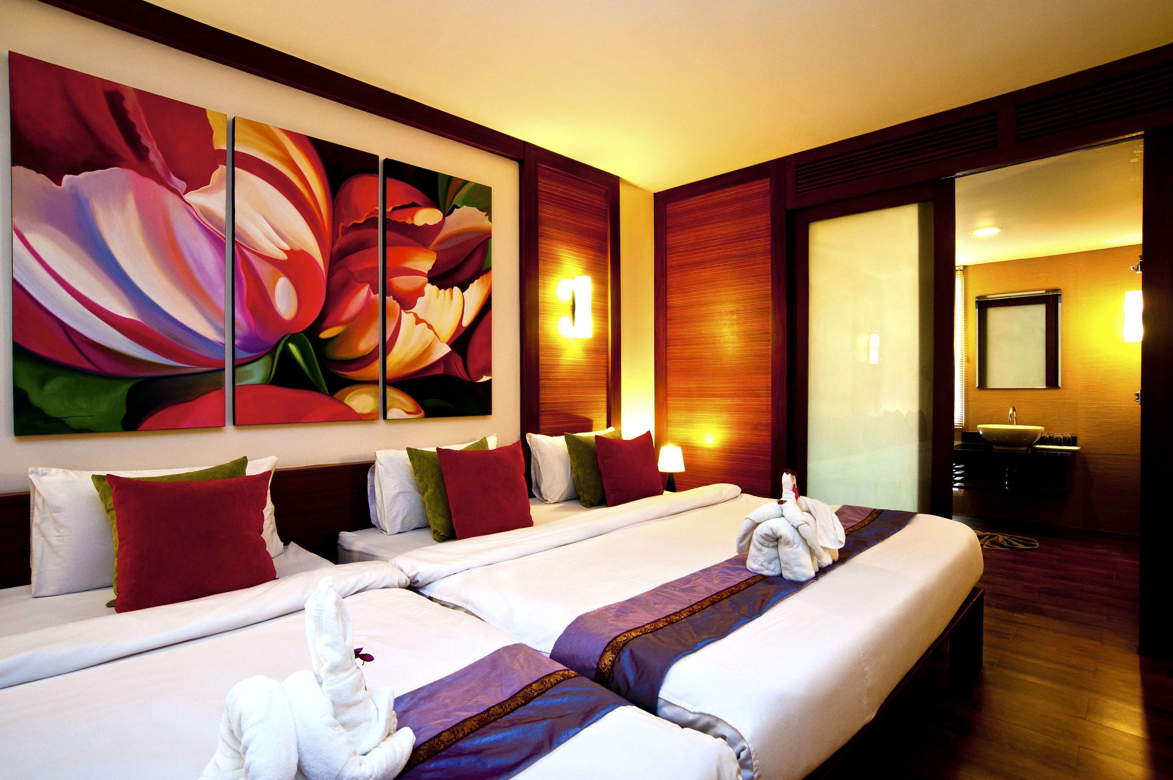 Habitación de hotel con dos camas, un cuadro grande con motivos florales, cabecero de madera, lámpara de noche y baño visible a través de una puerta de cristal.