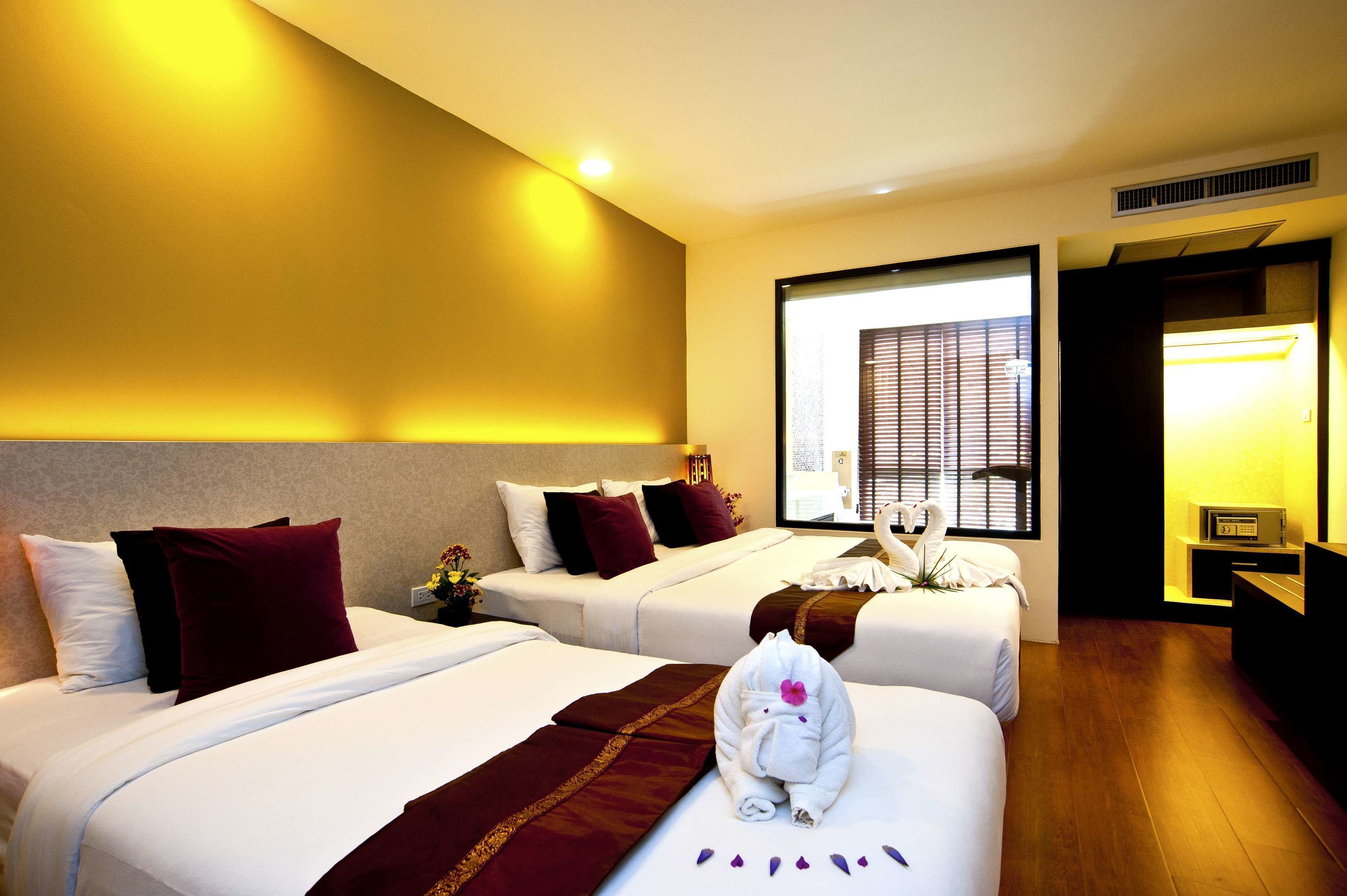 A hotel room with two beds, a large window with blinds, a bedside table with a lamp, and a TV.