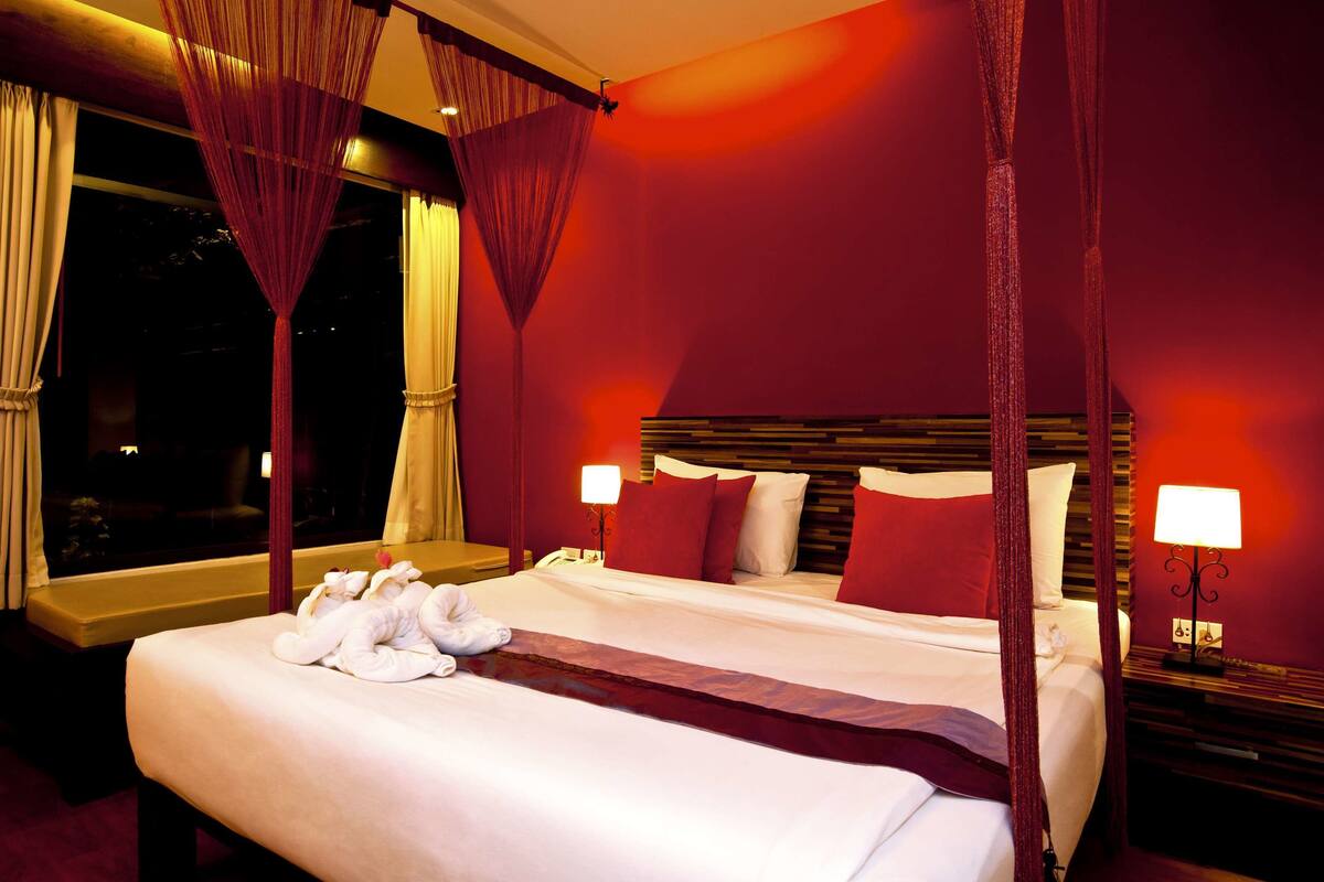 A hotel room with a large bed, red curtains, and a night view through the window.