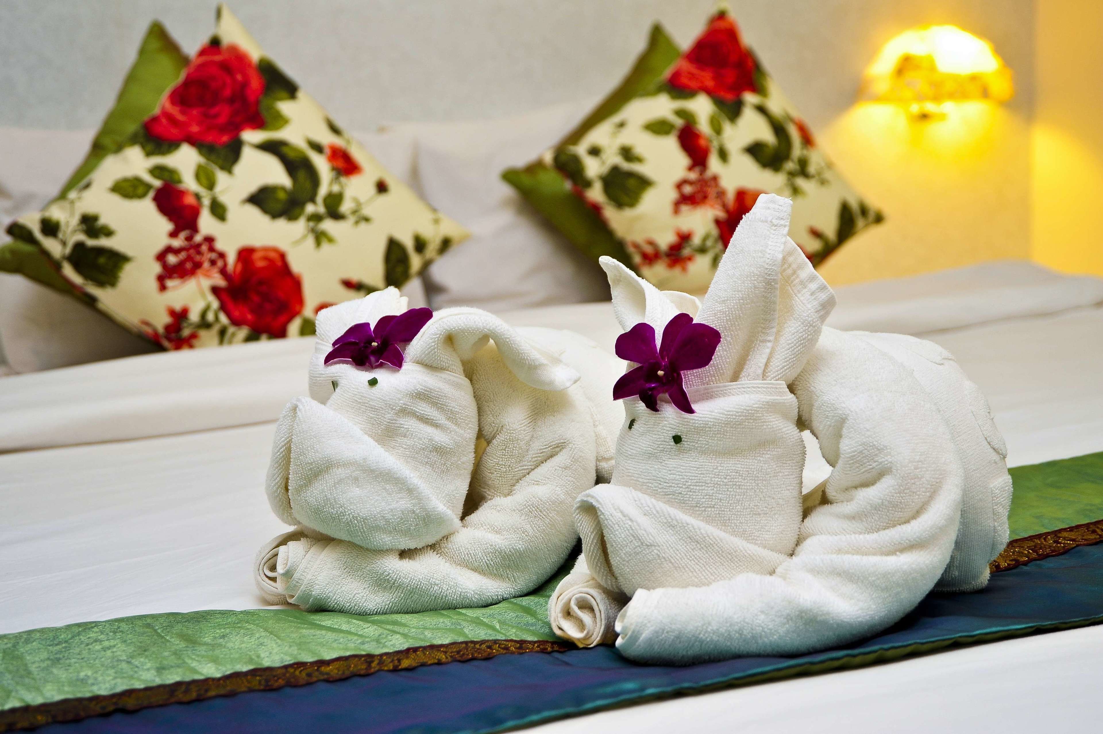 Zwei Handtücher sind dekorativ auf einem Bett drapiert, ergänzt durch blumige Kissen und eine Nachttischlampe.