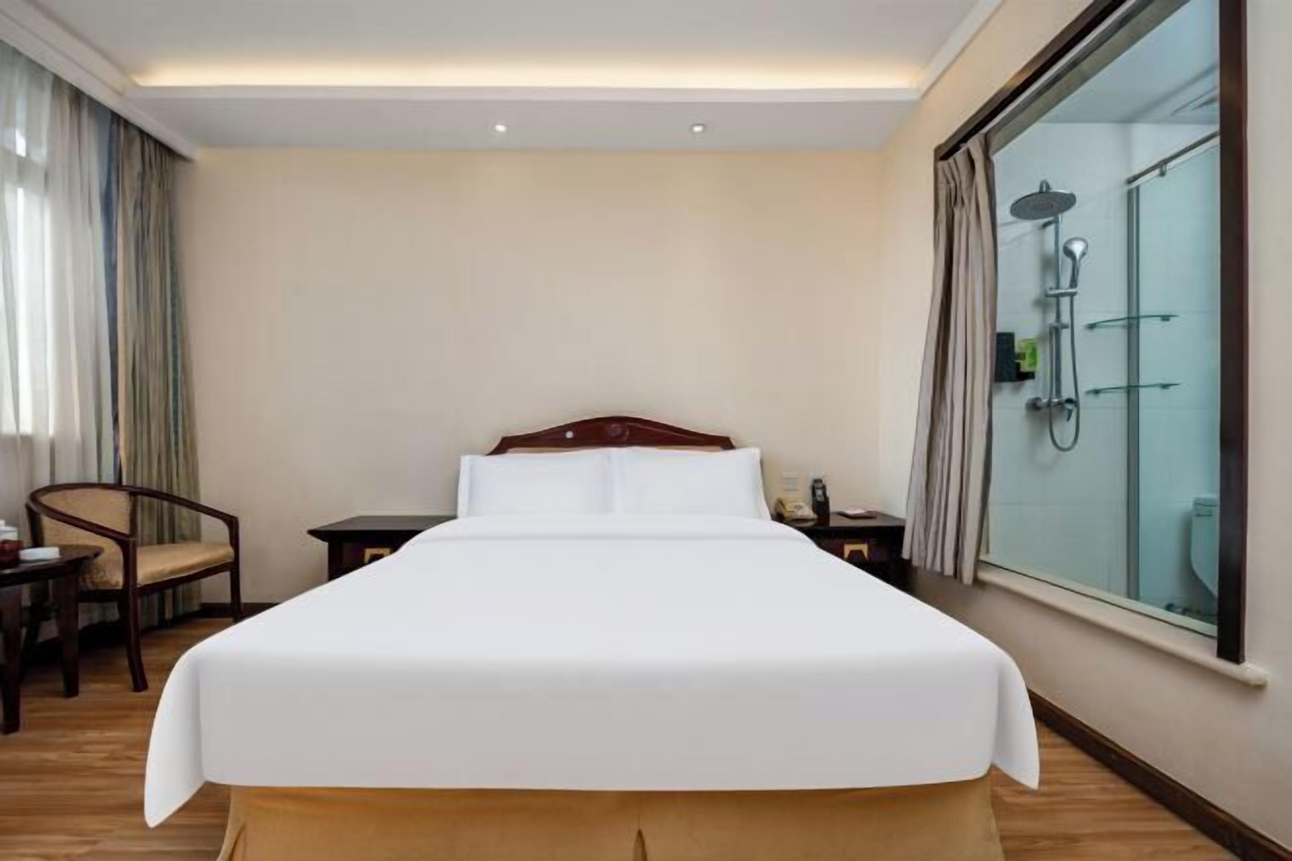 A hotel room with a bed, a chair, a small table, a nightstand, and a shower area.