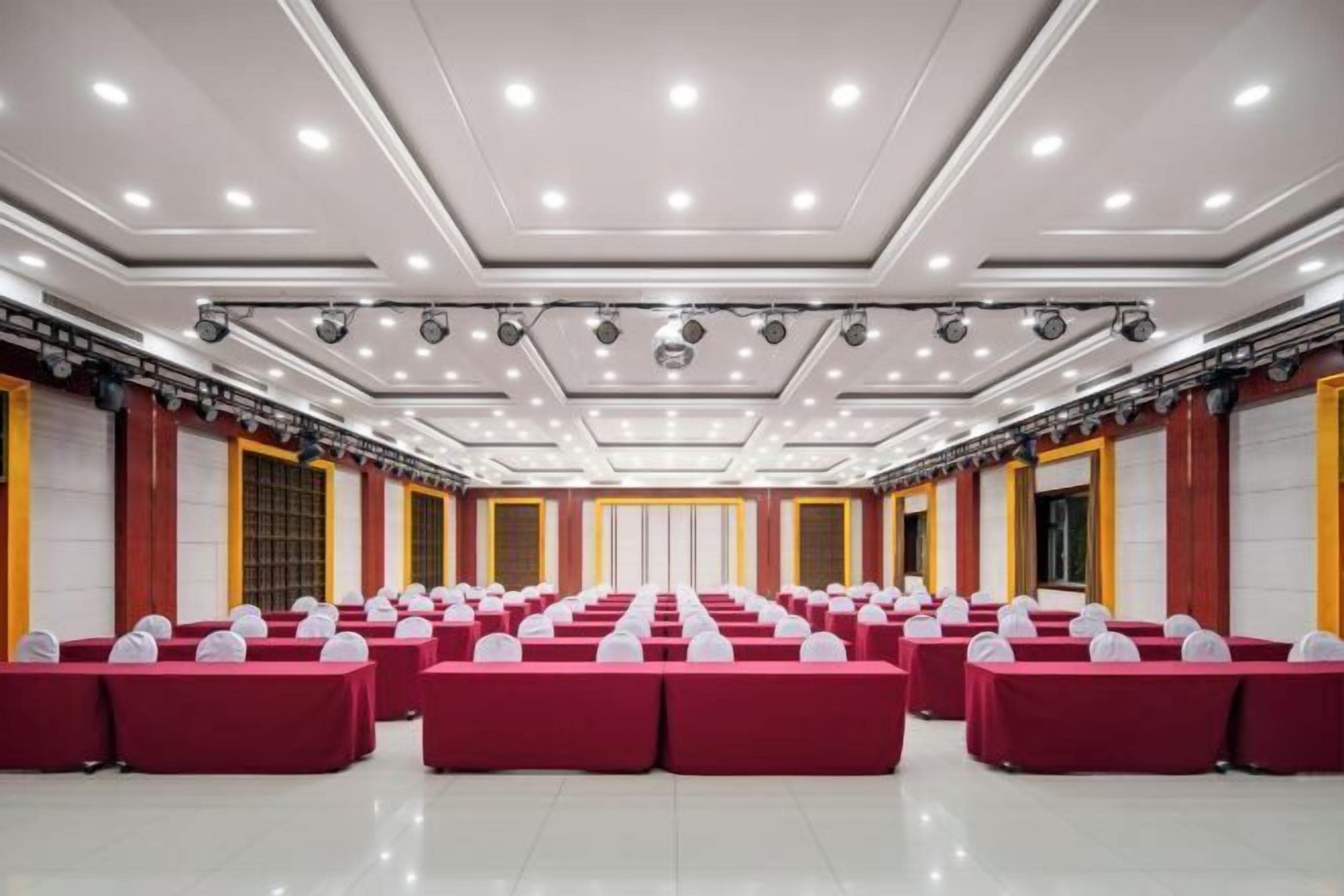 Sala de conferência com fileiras de mesas vermelhas e cadeiras brancas, equipada com luminárias no teto e sistema de som.