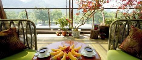 Una terraza con mobiliario de mimbre, una mesa con frutas y bebidas, y vistas a montañas.
