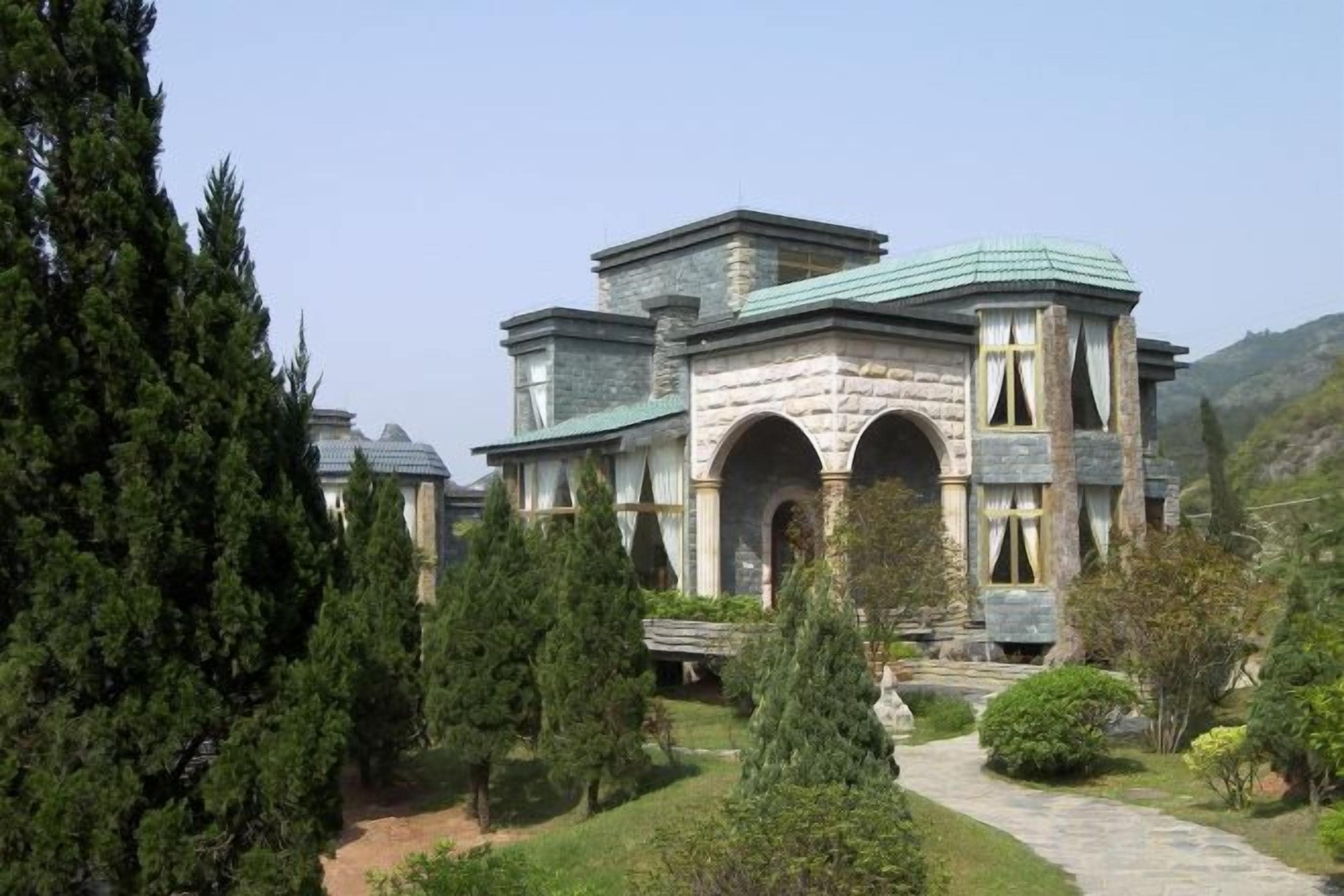 Un bâtiment en pierre doté de plusieurs arcades et d’un toit végétalisé, entouré d’arbres et bordé d’un sentier.